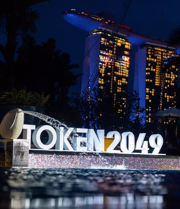 Illuminated 'TOKEN2049' sign with water feature in foreground and Marina Bay Sands hotel lit up at night in the background.